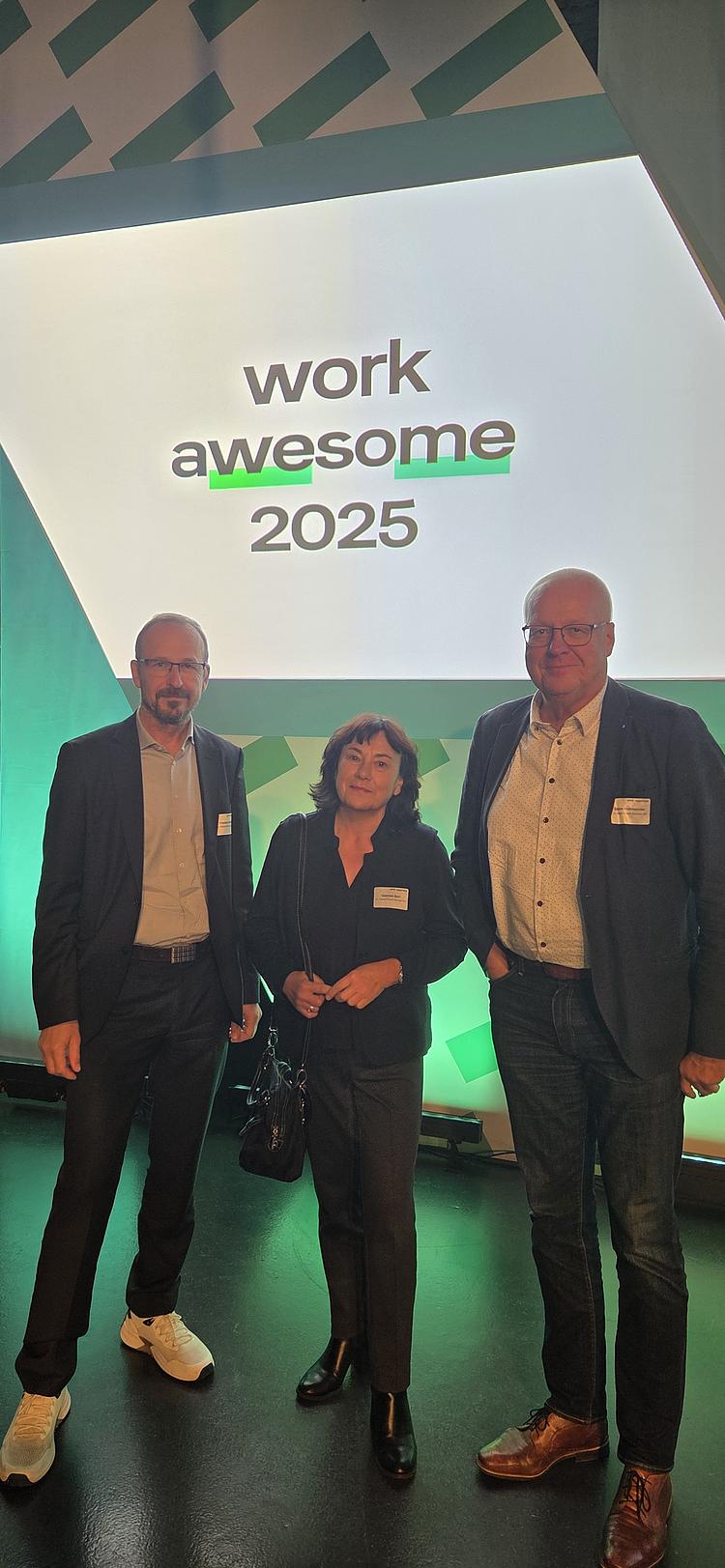 Vertreterinnen und Vertreter der Eckert Schulen nehmen an der Konferenz Work Awesome 2025 teil. Die Veranstaltung widmet sich den Zukunftsthemen moderner Arbeitswelten – von digitaler Transformation über Führungskultur bis hin zu neuen Lernformen. Drei Personen stehen vor einer Bühne mit der Aufschrift „Work Awesome 2025“.