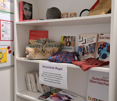 Fairschenkregal Weißes Fairschenkregal. Man sieht nur zwei bis drei Ablagen. Dort befinden sich Bücher, Dekogegenstände, CDs etc. zu Mitnehmen bereit.