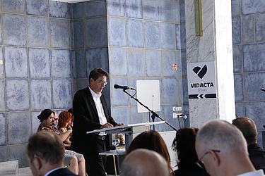 Worte, die bewegen Ein Mann im schwarzen Anzug steht an einem Rednerpult mit Mikrofon in einem Saal mit blauen Kacheln. Im Hintergrund sitzen Zuhörende. Rechts ist ein Schild mit der Aufschrift „VIDACTA Schulen“.
