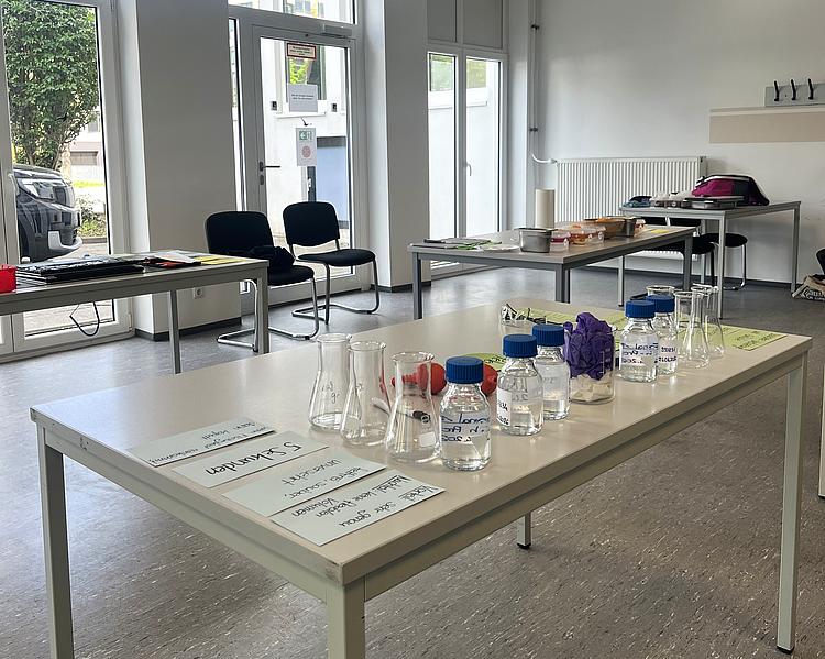 Laborraum mit mehreren Tischen, auf denen Glasgefäße, beschriftete Probenflaschen und Arbeitsmaterialien für ein Experiment vorbereitet sind.