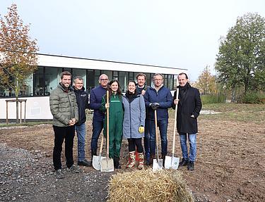 Eine Gruppe von acht Personen mit Schaufeln steht bei der Baumpflanzaktion am Campus der Eckert Schulen auf einem vorbereiteten Feld.
