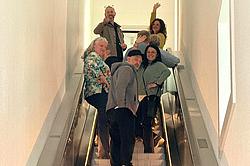 Teilnehmende der Mediengestalter-Exkursion fahren mit einer Rolltreppe durch den hell gestalteten Innenbereich des Museums und blicken in die Kamera.