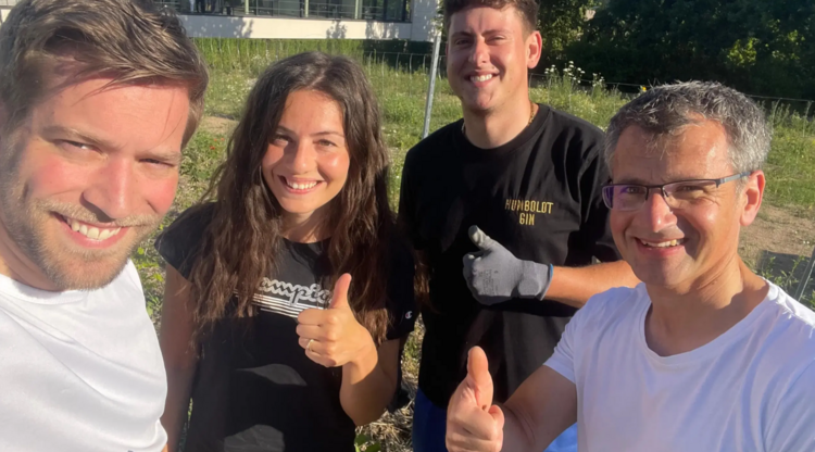 Vier Mitglieder des Projektteams posieren lachend und mit hochgestrecktem Daumen für ein Selfie auf der Fläche des Mikrowalds. Die Sonne scheint, im Hintergrund ist die Grünfläche zu erkennen.