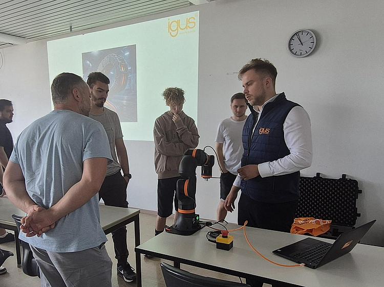 IGUS 2 Ein igus-Mitarbeiter demonstriert vor Schülern einen schwarzen Roboterarm auf einem Tisch, im Hintergrund eine Präsentation.
