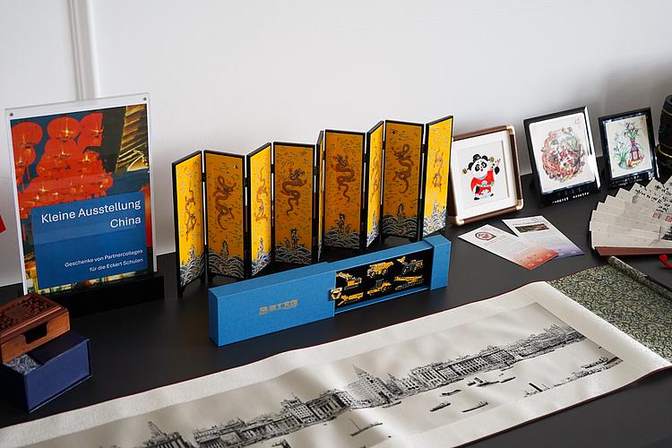 Auf einem Tisch sind verschiedene chinesische Schaustücke ausgestellt, darunter ein dekorativer Paravent, gerahmte Motive, Schriftstücke, ein Modellfahrzeug und weitere Geschenke chinesischer Partnercolleges.