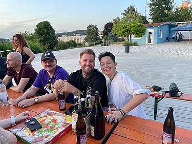 Eine Gruppe von Kolleg:innen sitzt an einem Holztisch im Freien mit Getränken und Pizza. Im Hintergrund sind Sandfläche, Bäume und ein Gebäude zu sehen. Alle wirken gut gelaunt.