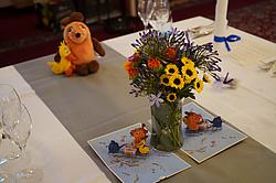 Blumenstrauß aus gelben, lila und orangefarbenen Blüten in einer Glasvase auf einem Tisch, dekoriert mit Geburtstagskarten und Stofffiguren der „Sendung mit der Maus“.