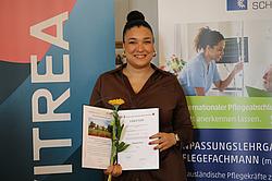 Eine Frau steht vor einem Banner, hält ihre Urkunde und eine Blume und lächelt in die Kamera.