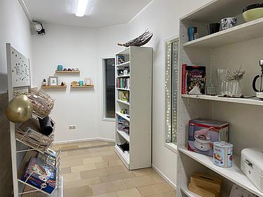 Das Bild zeigt einen kleinen, hellen Raum mit weißen Wänden und Regalen. Rechts stehen mehrere weiße Bücherregale mit verschiedenen Gegenständen: Kochbücher, Tassen, Gläser, eine Kaffeemaschine, eine Blechdose mit „Donuts“-Motiv und eine Box für einen Donut-Maker. Links sind offene Ablagen mit Spielen, darunter „Yakari“, sowie Körbe für Yoga- oder Sportmatten. An der Rückwand hängen zwei kleine Holzregale mit Dekoration wie Kerzen, Bilderrahmen und einer Figur. Der Raum wirkt wie ein Gemeinschafts- oder Aufenthaltsbereich für Freizeit und Entspannung.