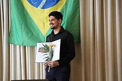 Ein Teilnehmer steht vor einer brasilianischen Flagge, hält seine Urkunde und eine Blume und lächelt stolz.