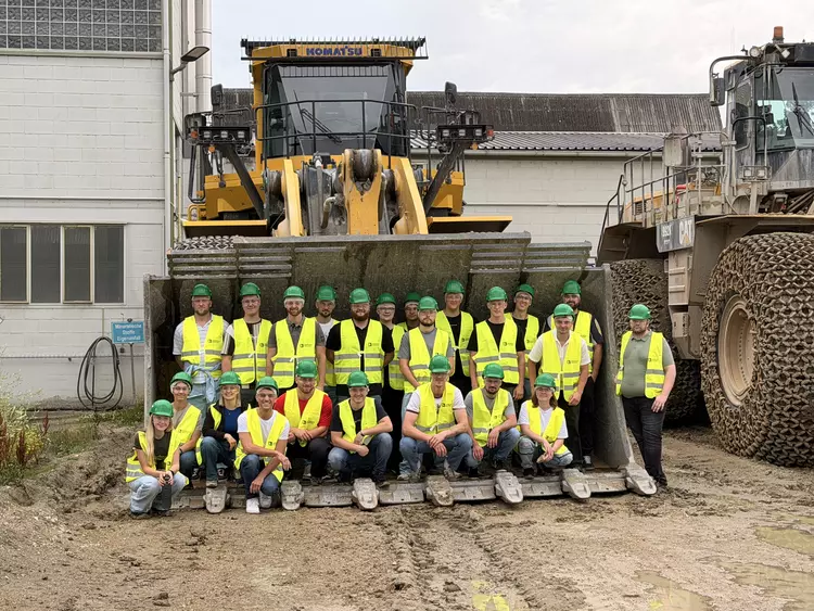 Gruppenfoto der Bautechnikerklassen der Eckert Schulen in gelben Warnwesten und grünen Schutzhelmen, aufgenommen in der Schaufel eines großen Radladers vor einem Werksgebäude von Heidelberg Materials.