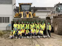 Gruppenfoto der Bautechnikerklassen der Eckert Schulen in gelben Warnwesten und grünen Schutzhelmen, aufgenommen in der Schaufel eines großen Radladers vor einem Werksgebäude von Heidelberg Materials.