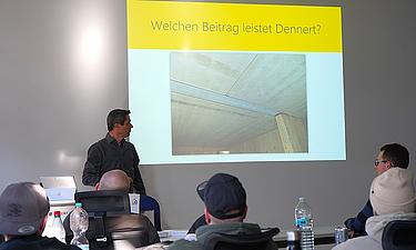 Herr Stangl von der Firma Dennert während eines Fachvortrags. Teilnehmende sehen die Folie mit dem Titel "Welchen Beitrag leistet Dennert"