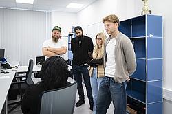 In einem Büro stehen vier Personen in einer kleinen Gruppe und schauen auf eine Frau, die mit dem Rücken zur Kamera in einem Bürostuhl sitzt. Die stehenden Personen wirken aufmerksam und hören der Dame zu.