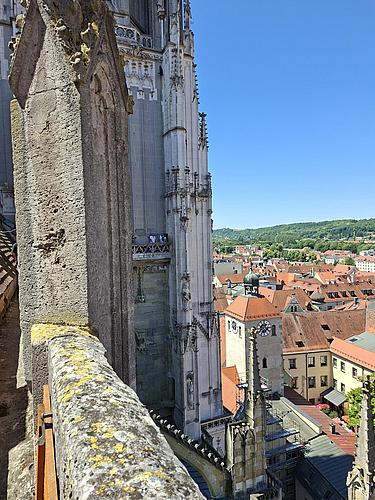 Von einer Balustrade aus fotografierter Blick auf die filigrane Fassade des Regensburger Doms mit ihren gotischen Details. Im Hintergrund erstrecken sich die roten Dächer der Altstadt und eine Turmuhr. Am Horizont ist eine grüne Hügellandschaft unter klarem blauen Himmel sichtbar.