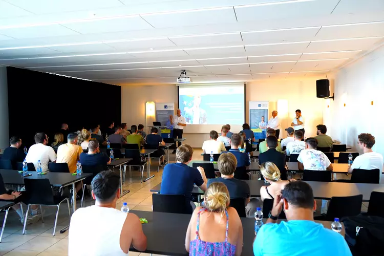 Willkommen zum Abenteuer Ausland Blick in einen Veranstaltungsraum mit zahlreichen Teilnehmenden, die einer Präsentation über das Auslandsprogramm der Eckert Schulen nach China im Herbst 2025 folgen. Im Hintergrund sprechen drei Männer vor einer Projektionsfläche.