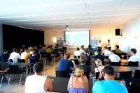 Willkommen zum Abenteuer Ausland Blick in einen Veranstaltungsraum mit zahlreichen Teilnehmenden, die einer Präsentation über das Auslandsprogramm der Eckert Schulen nach China im Herbst 2025 folgen. Im Hintergrund sprechen drei Männer vor einer Projektionsfläche.