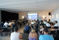 Willkommen zum Abenteuer Ausland Blick in einen Veranstaltungsraum mit zahlreichen Teilnehmenden, die einer Präsentation über das Auslandsprogramm der Eckert Schulen nach China im Herbst 2025 folgen. Im Hintergrund sprechen drei Männer vor einer Projektionsfläche.
