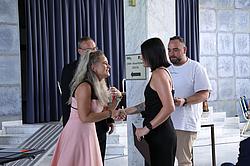 Eine Frau im rosa Kleid überreicht einer Absolventin in schwarzer Kleidung per Handschlag eine Urkunde. Im Hintergrund zwei weitere Personen, ein Mann hält ebenfalls eine Urkunde.