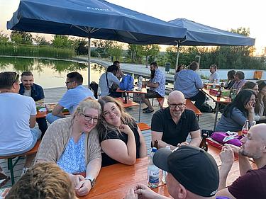 Mehrere Personen sitzen in Gruppen an Biertischen am Seeufer des Eckert Beach; im Vordergrund zwei Frauen, die sich anlehnen und in die Kamera lächeln, umgeben von Gesprächen, Getränken und Pizza.