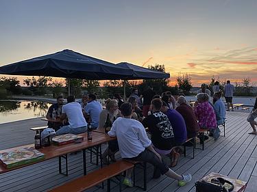 Ehemalige der MG-Klasse sitzen in Gruppen an Biertischen auf einem Holzdeck am Eckert Beach. Die Sonne geht malerisch hinter den Bäumen unter, das Licht spiegelt sich im Wasser. Pizzakartons und Getränke stehen auf den Tischen.