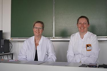Zwei Frauen in weißen Kitteln sitzen vor einer Tafel, lächeln in die Kamera. Eine trägt ein Namensschild mit der Aufschrift „Dr. Cornelia Börzl – Eckert Schulen“.