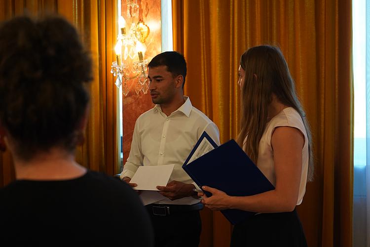 Zwei junge Erwachsene im Gespräch, beide halten Unterlagen in den Händen. Im Hintergrund goldene Vorhänge und Kronleuchter.