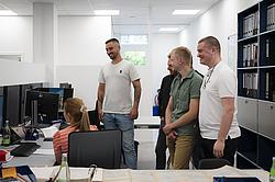 In einem modernen Büro stehen vier Männer lächelnd vor einer sitzenden Frau am Schreibtisch. Die Stimmung ist locker, die Gruppe ist begeistert von der Arbeit der Frau.