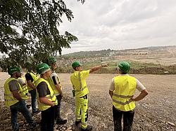 Studierende in Warnwesten und Schutzhelmen stehen am Rand des Kalksteinbruchs, während ein Mitarbeiter von Heidelberg Materials auf die gegenüberliegende Abbauwand zeigt und den Steinbruch erklärt.