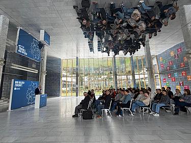 Teilnehmende sitzen in einem hellen Raum und hören einem Vortrag zu, der von vorne gehalten wird; eine spiegelnde Decke reflektiert die Szene.