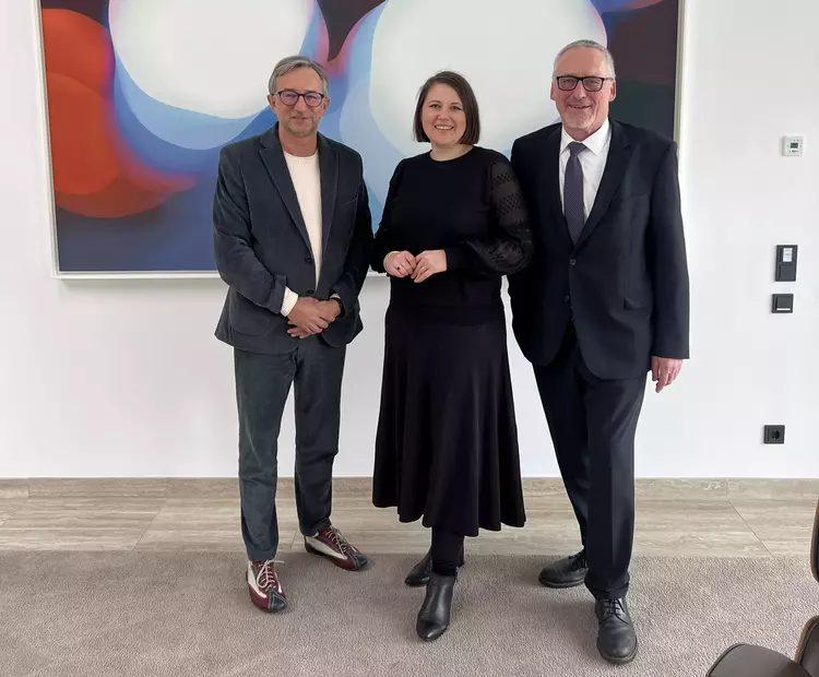 Drei Personen stehen vor einem abstrakten Kunstbild: Dr. Carolin Wagner in der Mitte, flankiert von Vorstand Stephan Koller und Geschäftsbereichsleiter Thomas Skowronek, beim Besuch der Eckert Schulen in Regenstauf.