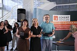 Zwei ausgezeichnete Absolvent:innen stehen mit einer Frau in der Mitte vor einem Roll-Up der Eckert Schulen. Sie halten Urkunden und Glastrophäen in der Hand, im Hintergrund stehen weitere Personen.