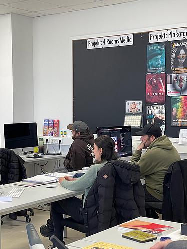 Unterrichtssituation mit mehreren Personen an Computern in einem Mediengestaltungsraum.