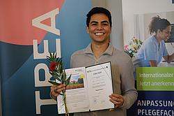 Ein Absolvent steht vor einem Informationsbanner und präsentiert stolz seine Urkunde und eine Blume.