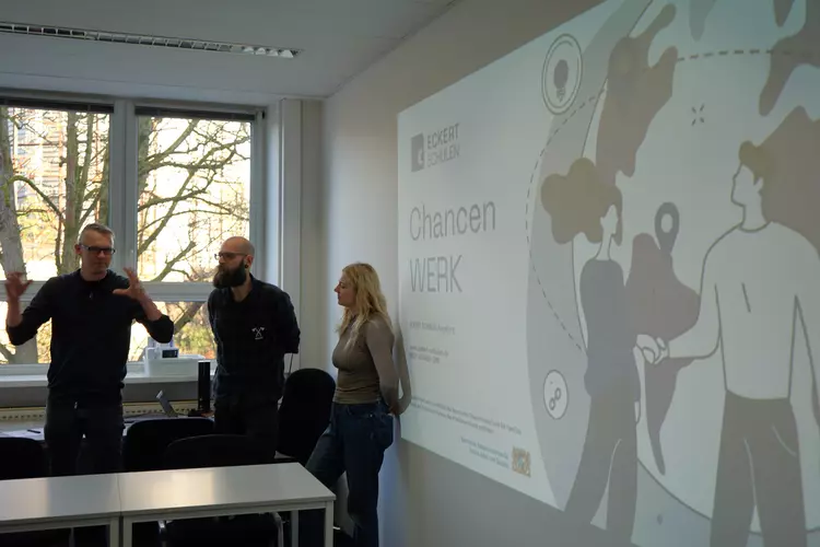 Drei Beteiligte stehen vor einer projizierten Präsentation des Projekts ChancenWerk an den Eckert Schulen und tauschen sich aus.