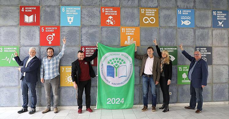 6 Personen stehen vor einer hellblau gefließten Wand. An der Wand befinden sich auf den Kacheln jeweils bunte Aufkleber mit den Nachhaltigkeitszielen in unterschiedlichen Farben z.B.: blau, rot, gelb, orange usw. Die Personen mittig halten gemeinsam ein Banner mit dem Aufdruck "Umweltschule" in grün hoch. Die Personen an den Rändern zeigen auf die Ziele an der Wand hinter ihnen. 