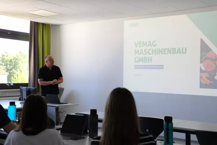 Thomas Dippel hält einen Vortrag vor angehenden Lebensmittelverarbeitungstechnikern. Auf der Präsentationsfolie ist die VEMAG Maschinenbau GmbH zu sehen.