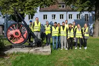 Gruppenfoto von elf Personen in gelben Warnwesten vor einem blauen Gebäude, links im Bild ein schwarzer Schiffsanker mit rotem Rad.