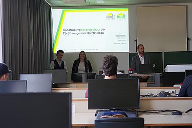Drei Personen präsentieren vor einer Projektionsfläche in einem Computerraum ihre Projektarbeit mit dem Titel „Konstruktiver Brandschutz der Türöffnungen im Holztafelbau“. Im Vordergrund sind Zuhörer an Computerarbeitsplätzen zu sehen.
