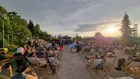 BBCO Eckert Beach Viele Besucher sitzen auf Liegestühlen am Sandstrand und blicken auf eine Bühne mit Musikern. Die Sonne geht im Hintergrund unter und taucht die Szene in warmes Licht.