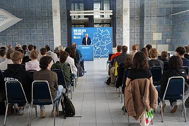 Ein Referent steht vor einem Publikum aus Jugendlichen und Erwachsenen und spricht an einem Rednerpult mit Eckert-Schulen-Branding über Bildungsangebote.