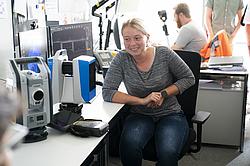 Eine junge Frau sitzt lächelnd an einem Schreibtisch in einem Büro. Vor ihr befinden sich verschiedene technische Geräte und Messinstrumente, darunter ein Laserscanner oder Tachymeter