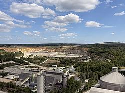 Blick von oben auf das Werksgelände von Heidelberg Materials in Burglengenfeld mit Produktionshallen, Silos und dem angrenzenden Kalksteinbruch, umgeben von Wald und Feldern.
