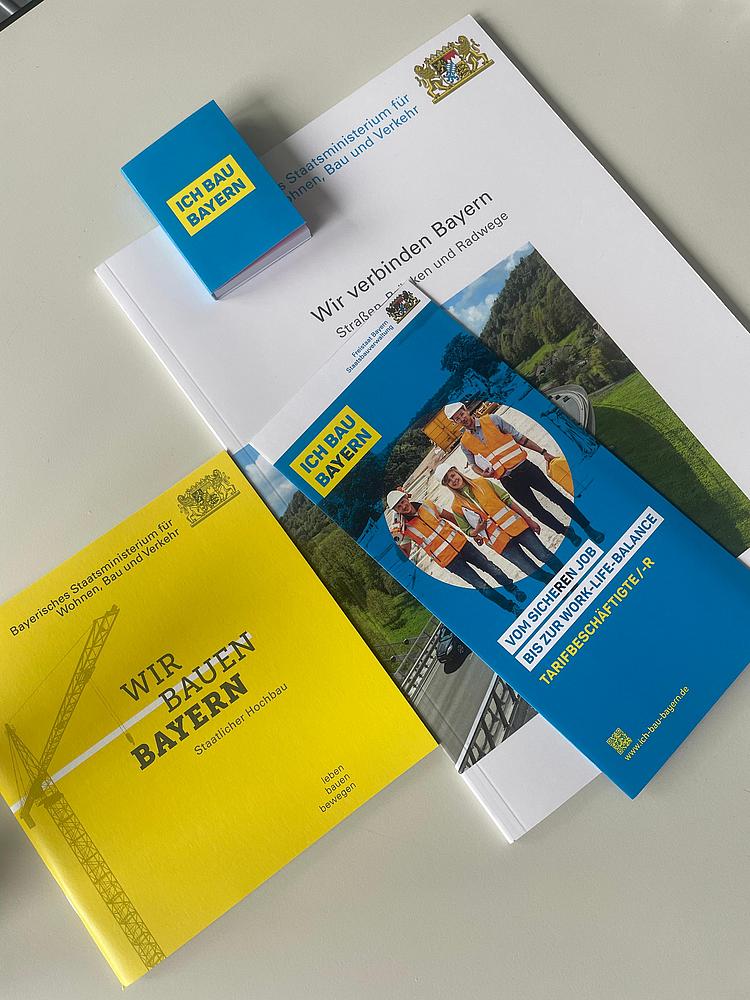 Auf einem Tisch liegt eine Auswahl an Informationsmaterialien des Bayerischen Staatsministeriums für Wohnen, Bau und Verkehr. Zu sehen sind eine gelbe Broschüre mit dem Titel „Wir bauen Bayern“, ein blauer Flyer „Ich bau Bayern“ mit einem Foto von Bauarbeitern in orangenen Warnwesten, eine weiß-graue Broschüre „Wir verbinden Bayern“ sowie ein kleiner blauer Notizblock. Die Materialien informieren über Bauprojekte, Infrastruktur und Karrieremöglichkeiten im staatlichen Bauwesen.