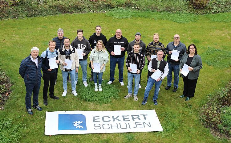 Gruppenfoto von oben von Energiebeauftragten der Dr. Eckert Akademie. Die Gruppe steht in einer grünen Wiese und wird von oben fotografiert. Vor der Gruppe liegt ein Banner mit dem Logo der Eckert Schulen auf dem Boden.