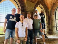 Acht Männer unterschiedlichen Alters stehen in einem Turm mit rotem Backstein und Spitzbogenfenstern. Durch die vergitterten Fenster fällt Licht ein. Die Gruppe lächelt in die Kamera, im Hintergrund sind Landschaft und Himmel zu erkennen.