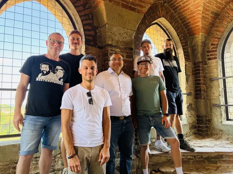 Acht Männer unterschiedlichen Alters stehen in einem Turm mit rotem Backstein und Spitzbogenfenstern. Durch die vergitterten Fenster fällt Licht ein. Die Gruppe lächelt in die Kamera, im Hintergrund sind Landschaft und Himmel zu erkennen.