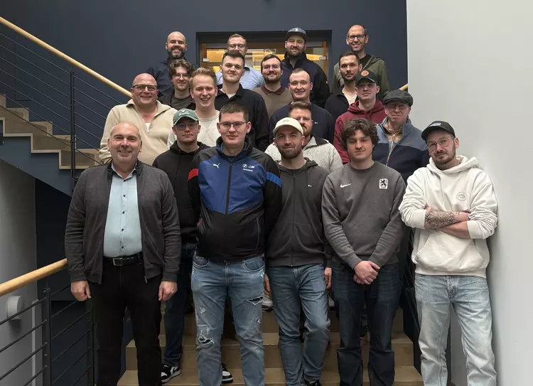 Eine größere Gruppe von Männern, bestehend aus Studierenden und Unternehmensvertretern, steht gestaffelt auf einer Treppe in einem Firmengebäude und posiert für ein gemeinsames Gruppenfoto.