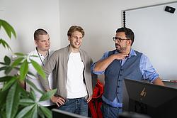 Drei Männer stehen in einem Büro vor einem Monitor und unterhalten sich. Zwei Männer lächeln, das Gespräch wirkt locker und entspannt.