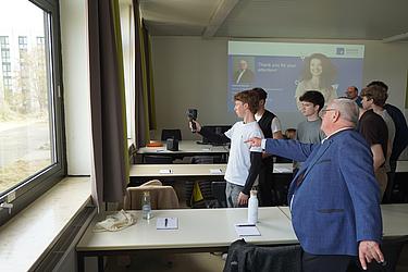 Ein Schüler misst mit einer Wärmebildkamera an einem Fenster, während ein Dozent unterstützend daneben steht.
